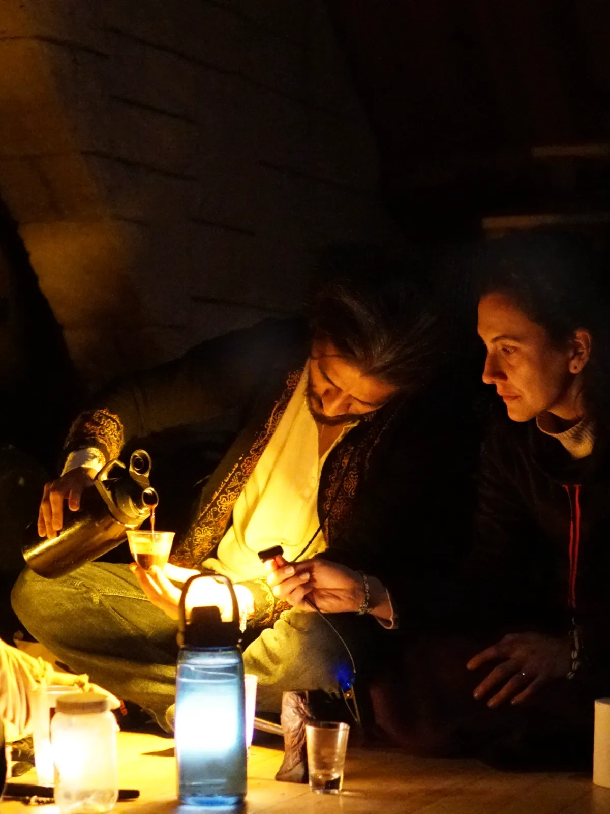 Dos personas sentadas en el piso sirven un vaso a la luz de una lámpara, de noche