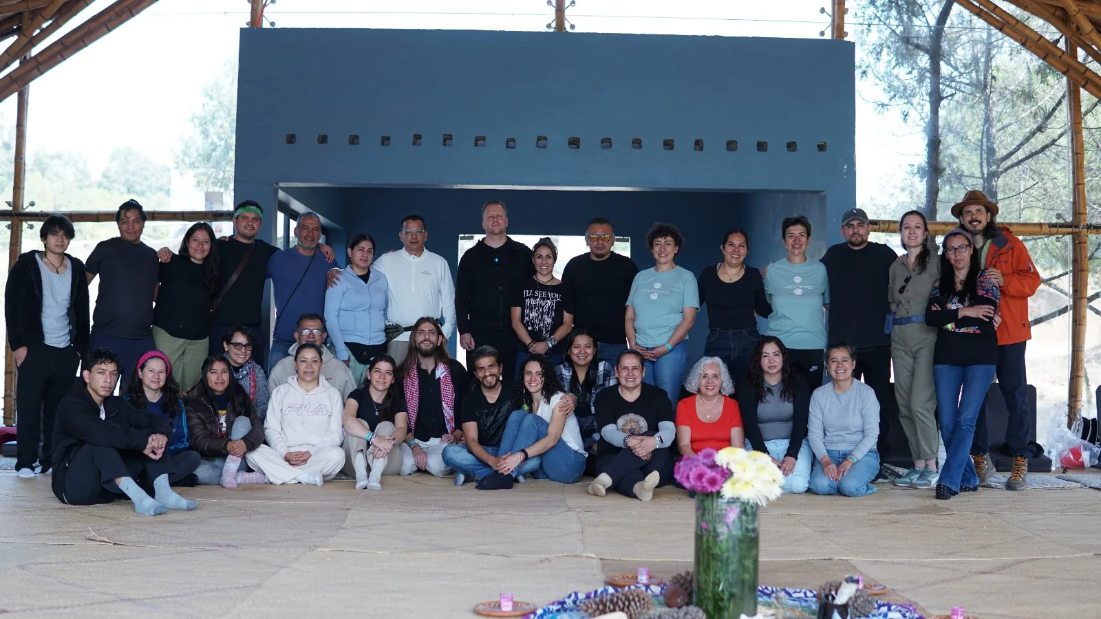 Grupo de participantes reunido bajo la palapa de bambú, de pie y sentado sobre el petate